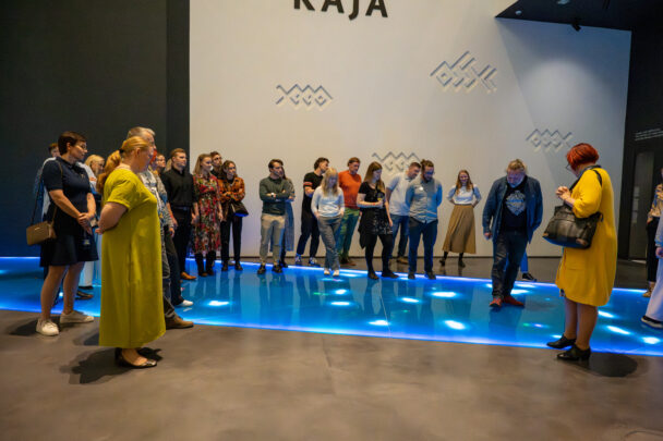 Umbes kahekümne inimesega grupp seisab galeriiruumi sinise valgustusega platvormi ümber; taustal valge sein