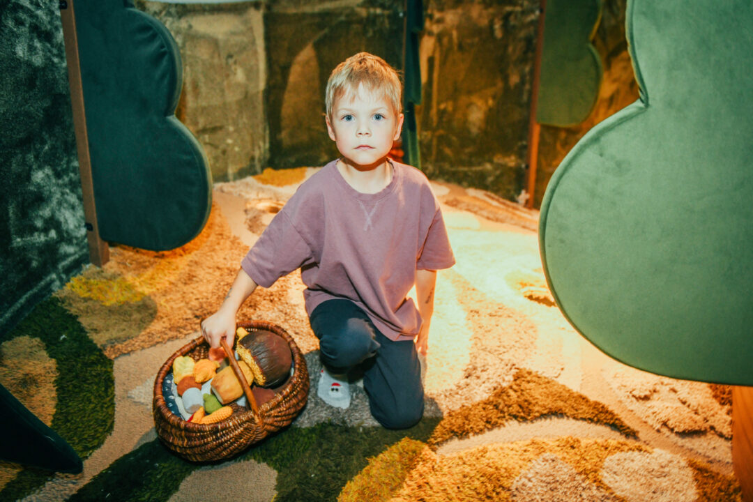 Väike poiss kükitab metsa meenutaval vaibal ja hoiab paremas käes puidust punutud korvi. Korv on täis värvilisi pehmeid mänguasju.